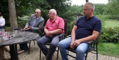 Trzech sołtysów siedzi przy stoliku zewnętrznym na tle roślinności 
